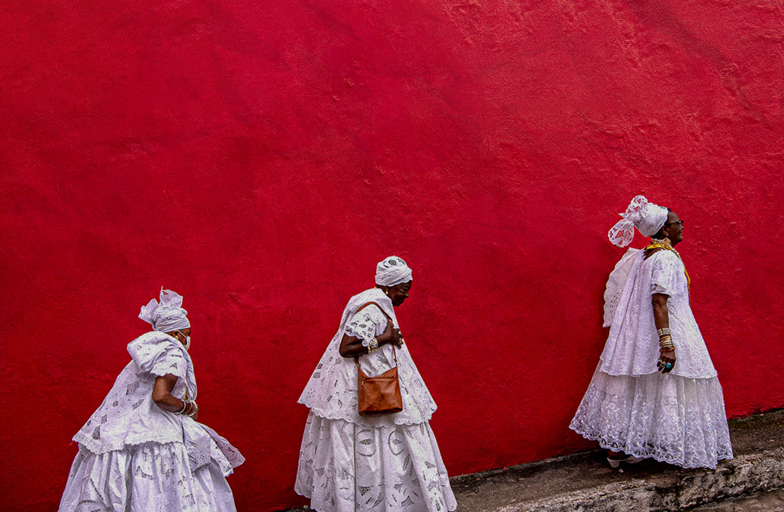 Milagres do POVO — mulheres com trajes de baiana caminhando em frente a uma parede vermelha