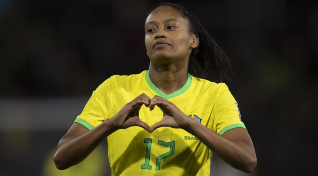A jogadora Ary Borges fazendo um coração com as mãos. Ela tem a pele negra e os cabelos longos preso em um rabo de cavalo. A jogadora veste a camisa da Seleção Brasileira, em amarelo e verde, com o número 17.