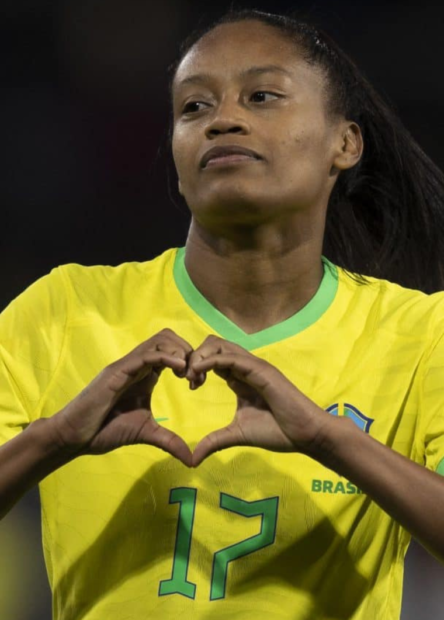 A jogadora Ary Borges fazendo um coração com as mãos. Ela tem a pele negra e os cabelos longos preso em um rabo de cavalo. A jogadora veste a camisa da Seleção Brasileira, em amarelo e verde, com o número 17.